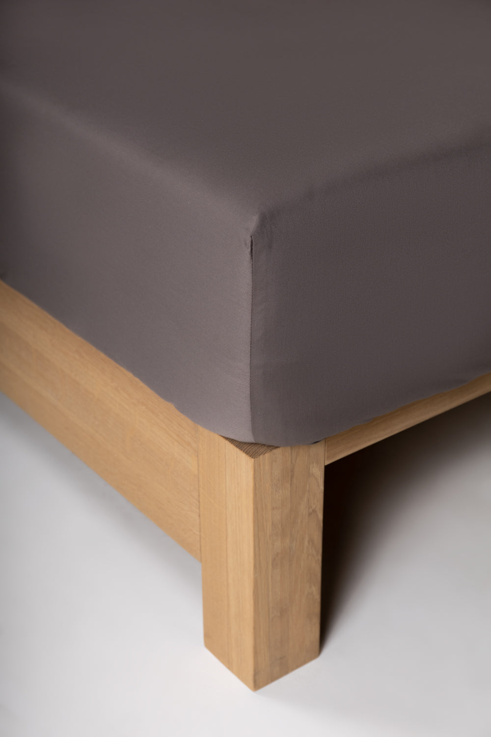 A corner of a gray fitted sheet on a bed, showing the rubber strip on the corner and the wood legs of the bed frame.